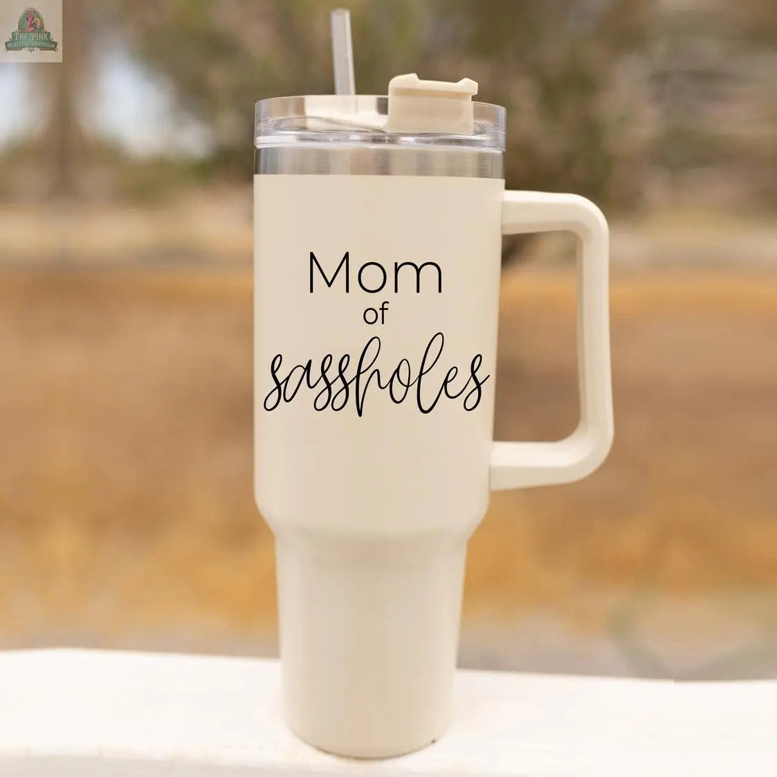 The Sassholes 40oz mug, a large cream-colored insulated travel mug with handle and straw, displays "Mom of assholes" in black cursive and print. Made by Gia Roma, it’s shown outdoors with a blurred background featuring trees.
