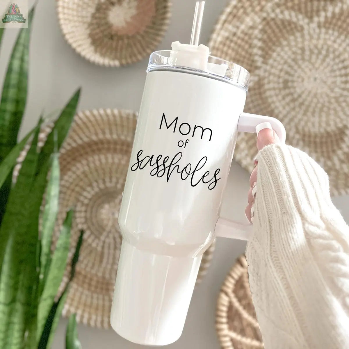 A hand in a cozy cream sweater holds the Sassholes 40oz insulated mug with a straw, featuring the phrase “Mom of sassholes.” Woven baskets and a green plant are softly blurred in the background.