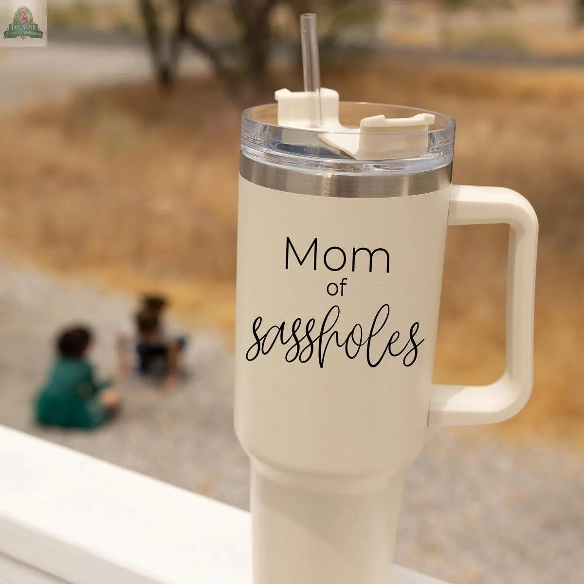 The Sassholes 40oz mug is a large cream-colored insulated tumbler with a straw, featuring the text "Mom of sassholes." Two children can be seen sitting on a dirt path outdoors in the blurry background.