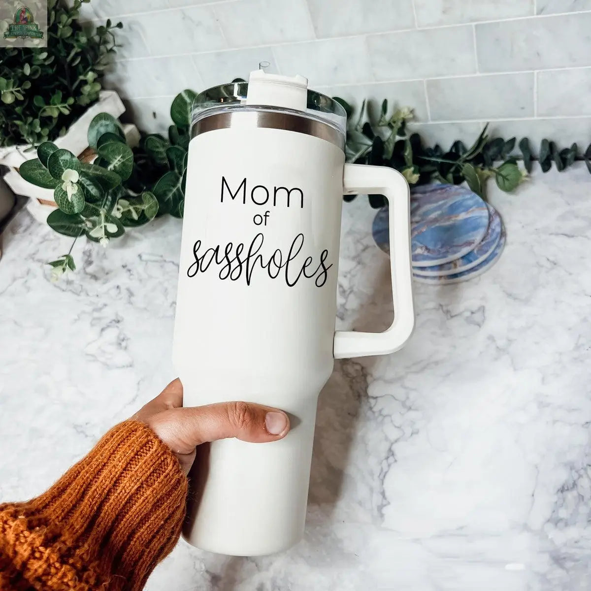 A hand holds the Sassholes 40oz insulated mug by Gia Roma with "Mom of sassholes" printed on it. The large white travel mug rests on a marble countertop, surrounded by green plants and round coasters.