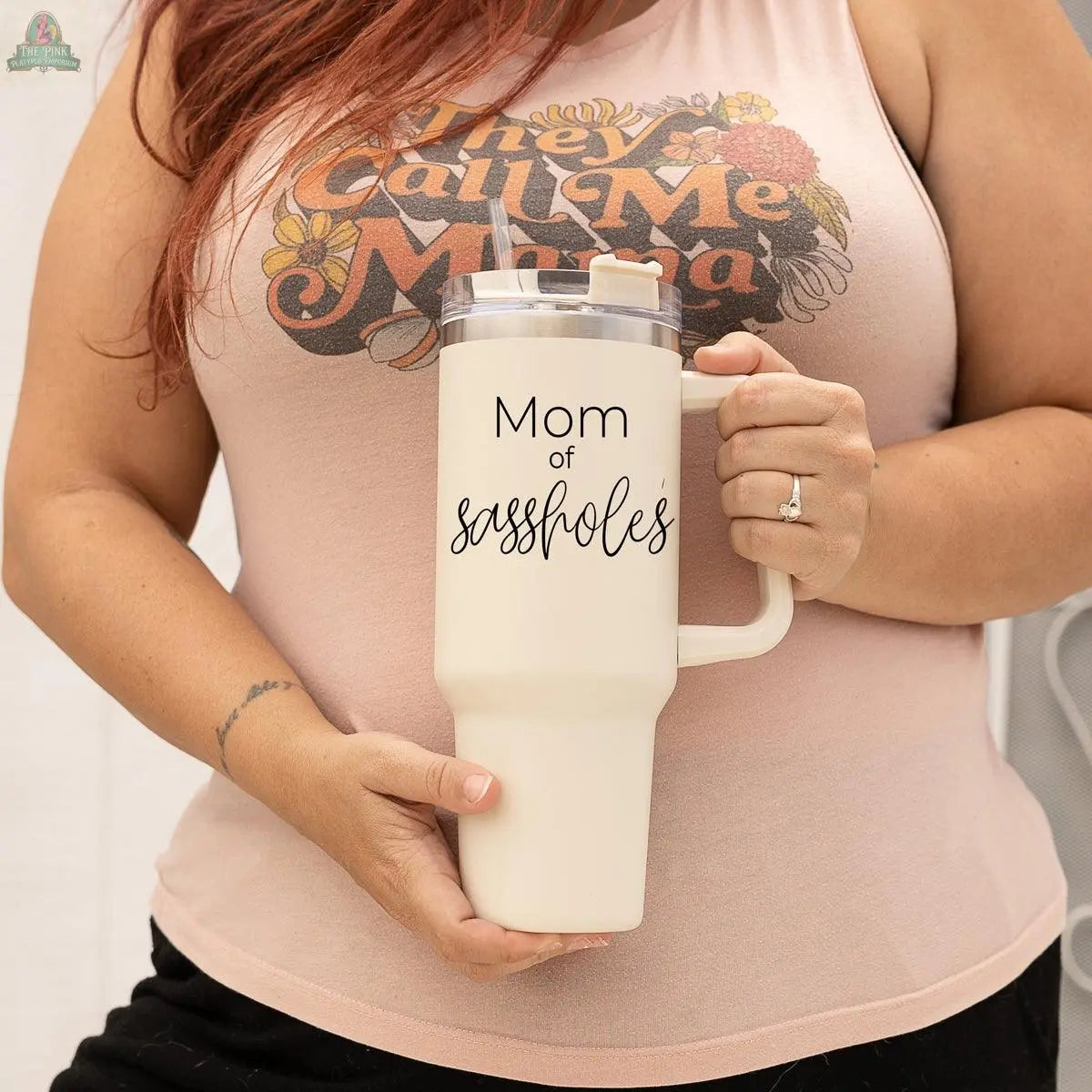 A woman wearing a sleeveless "They Call Me Mama" shirt holds a beige 40oz Sassholes travel mug by Gia Roma that reads "Mom of sassholes." She also has a ring on her finger.