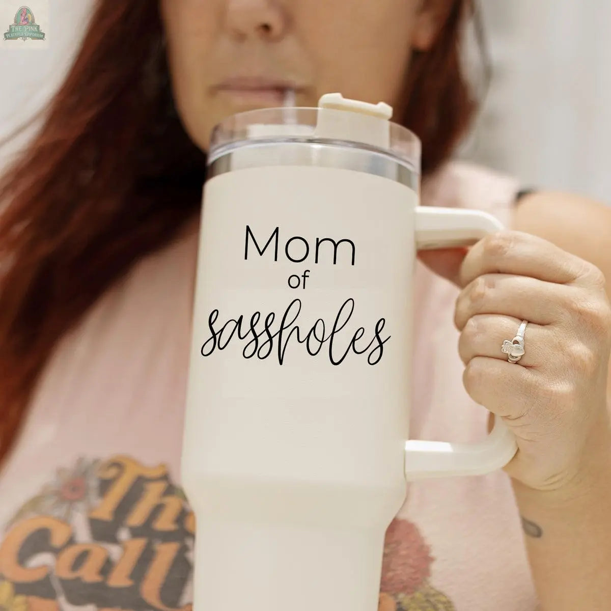 A woman holds the Sassholes 40oz travel mug, featuring "Mom of sassholes" text. She wears a Gia Roma graphic tee and a visible ring, with her face partially out of focus.