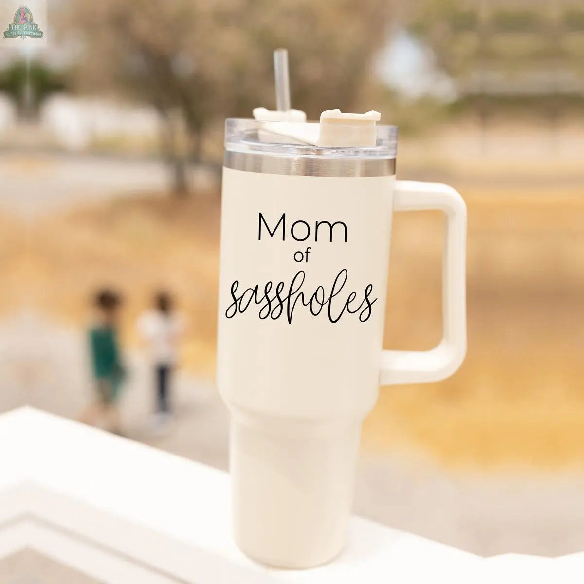 A large cream-colored Sassholes 40oz insulated mug with a handle by Gia Roma sits on a white railing, featuring the words "Mom of sassholes." Three children stand together in the blurred background outside.