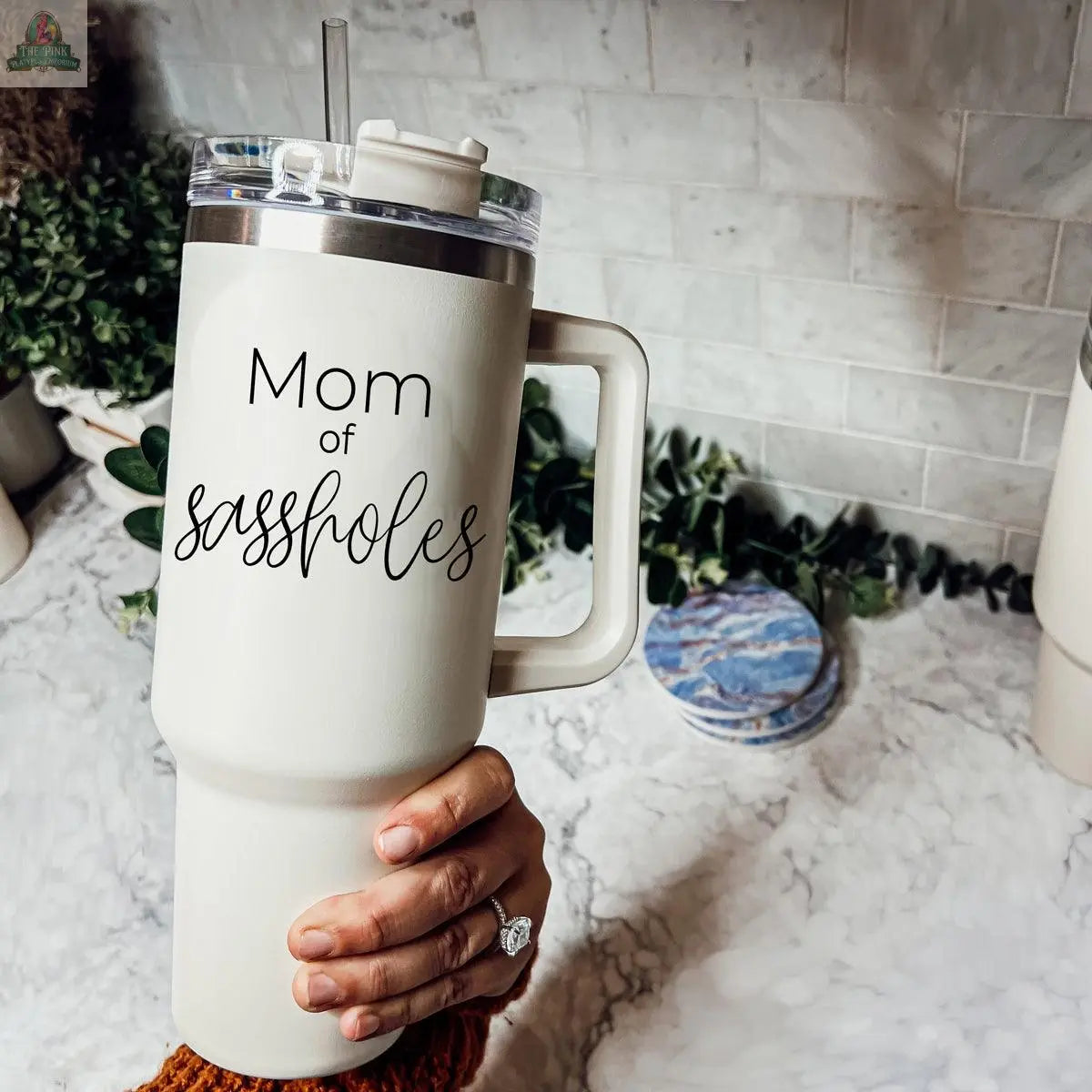 A hand holds a large Sassholes 40oz insulated travel mug with a lid and straw. The mug features a humorous phrase. A marble countertop, greenery, and white tiled wall are visible in the background.