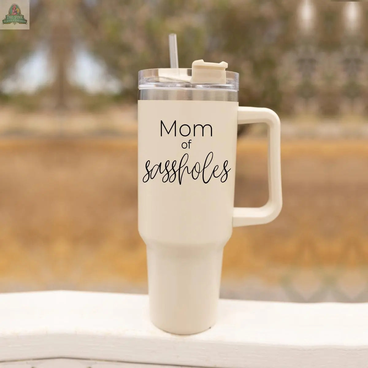 A beige Sassholes 40oz travel mug from Gia Roma, featuring "Mom of sassholes" in black script, with a handle and straw, sits on a ledge against a blurred outdoor background.