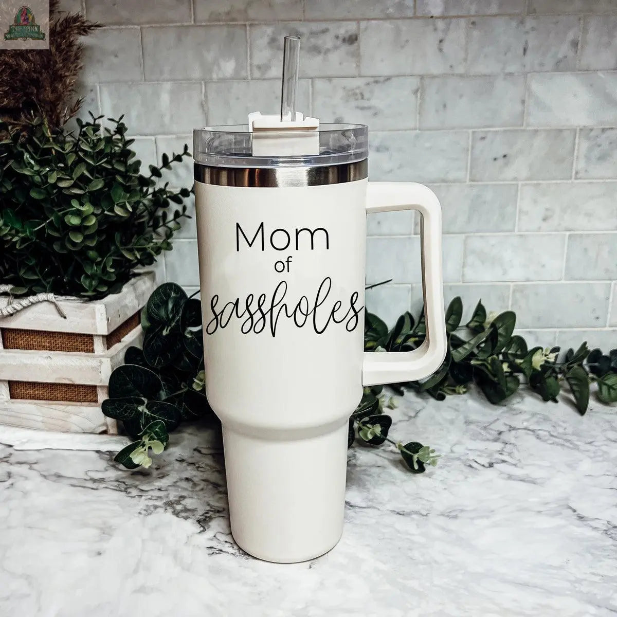 A Sassholes 40oz large white travel mug with a handle and straw sits on a marble countertop, labeled "Mom of sassholes" in black script. Green plants and white tiles make up the background.