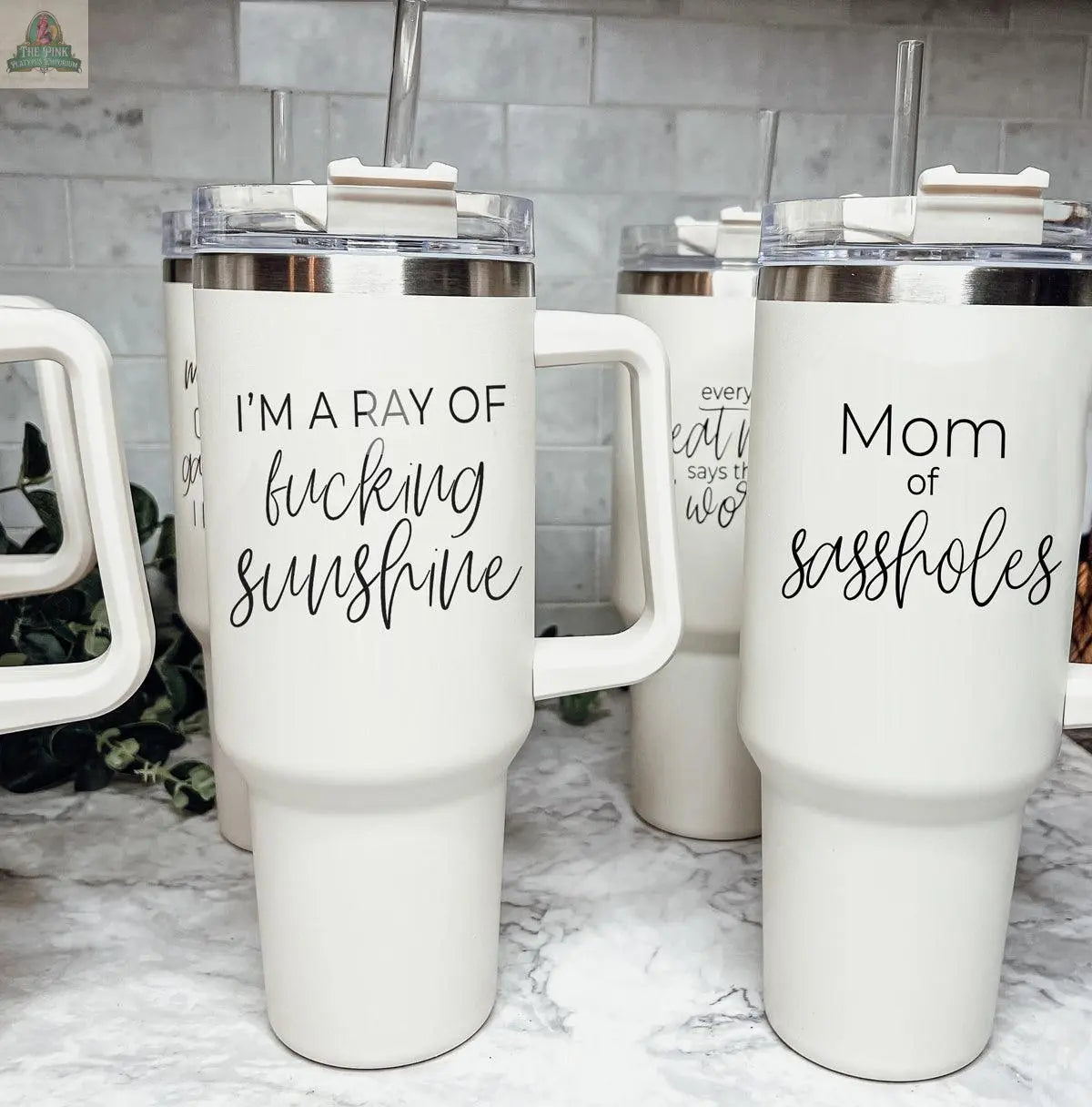 Four white Sassholes 40oz travel mugs feature funny, explicit quotes in black script, including "I’m a ray of fucking sunshine" and "Mom of sassholes," shown on a marble counter with a tiled backsplash.