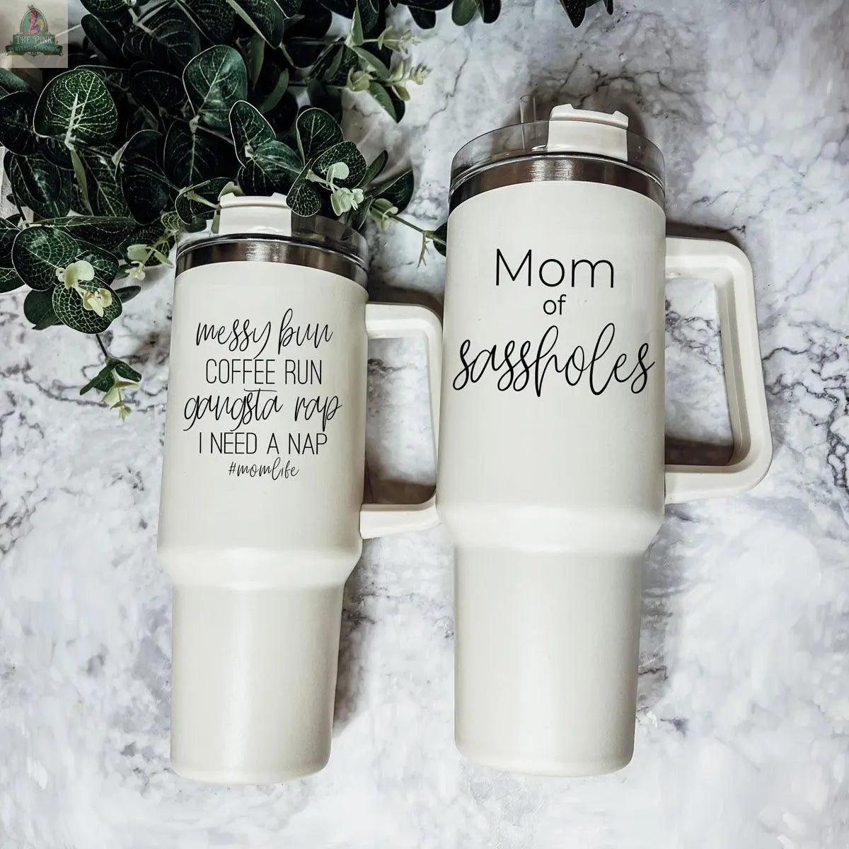Two white Sassholes 40oz insulated mugs with handles sit side by side. One reads “messy bun, coffee run, gangsta rap, I need a nap #momlife,” and the other says “Mom of sassholes.” Green plants are in the background.