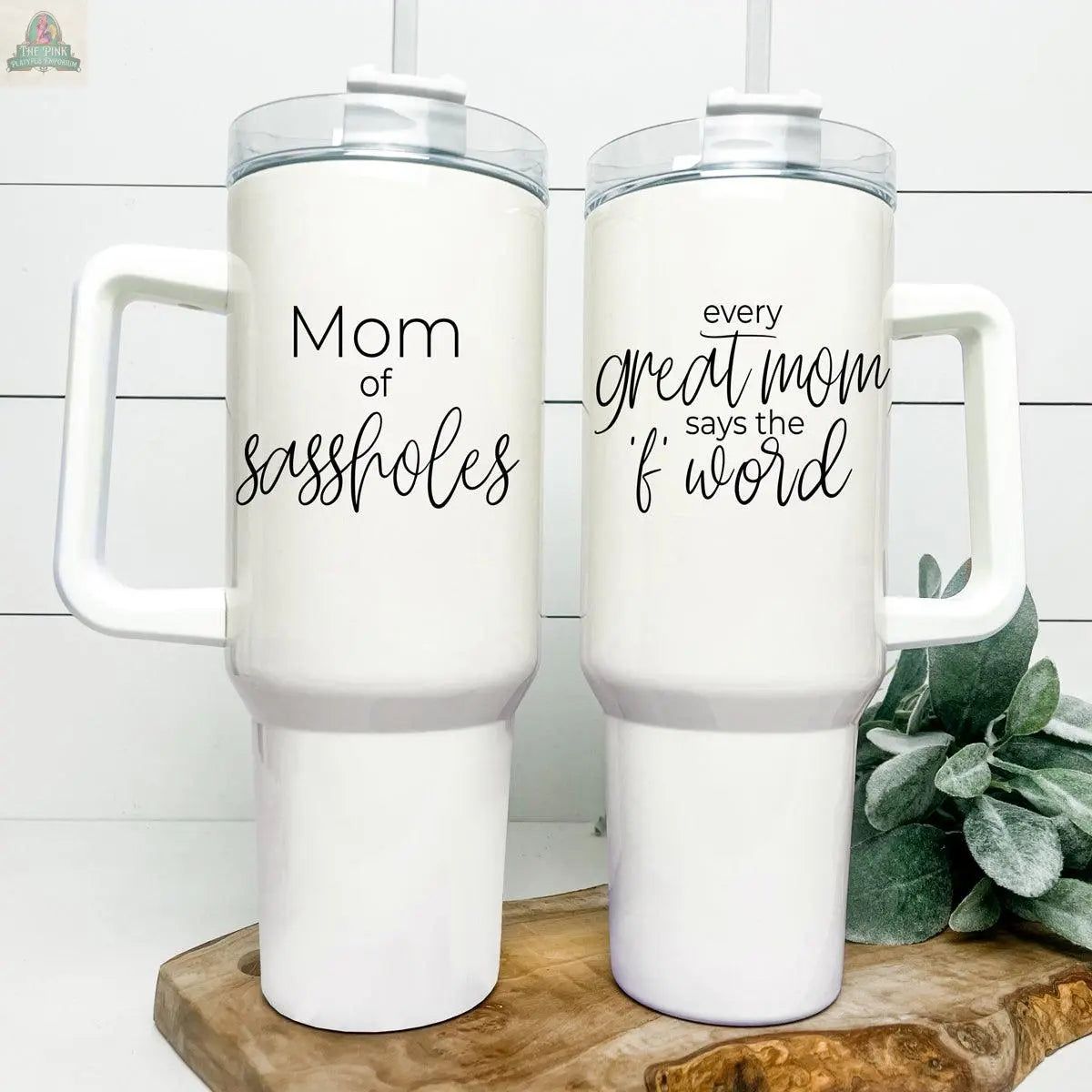 Two white Sassholes 40oz insulated mugs with handles and lids are displayed—one says "Mom of sassholes," the other "Every great mom says the f word." Both travel mugs rest on a wooden board with greenery beside them.