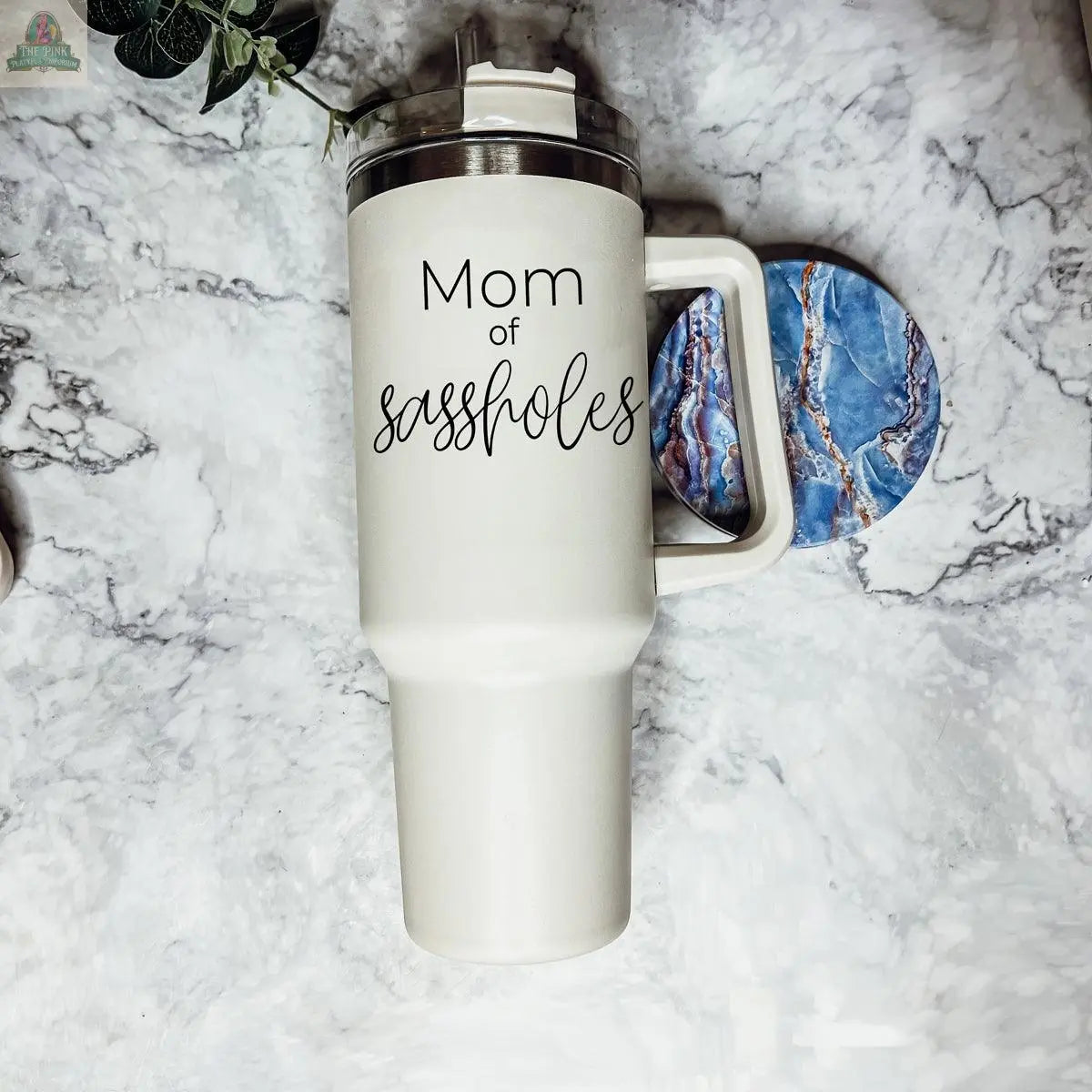 A tall white insulated Sassholes 40oz mug with handle and lid sits on marble, reading "Mom of sassholes" in black script. A blue marbled coaster is partly visible. This mug keeps drinks hot or cold in style.