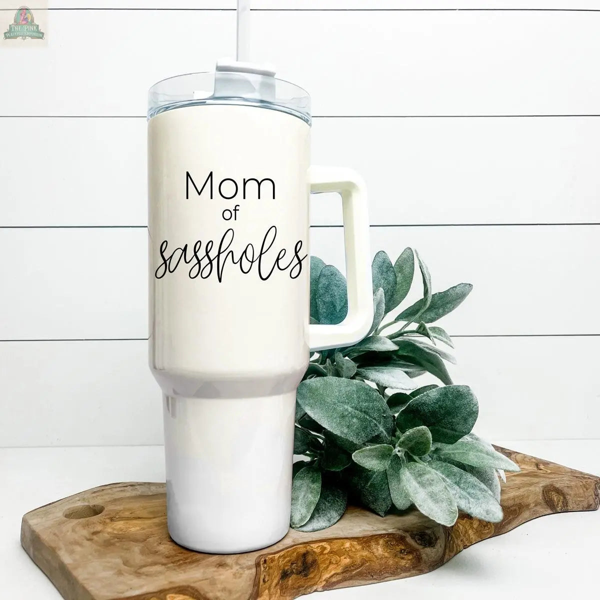 A tall white Sassholes 40oz insulated mug with handle and straw, featuring "Mom of sassholes," sits on a wooden board near green plants against a white shiplap backdrop. Keeps drinks hot or cold for hours.