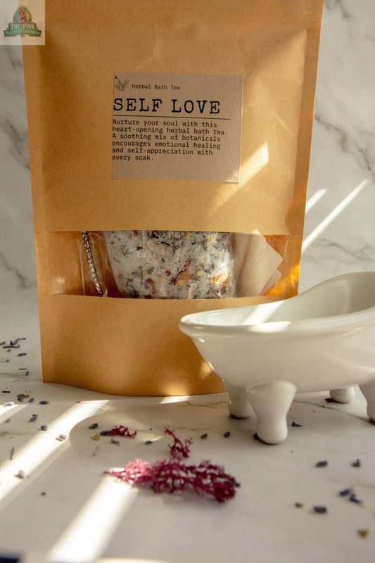 A brown paper pouch labeled Self Love herbal bath tea, infused with rose and vanilla essential oils, sits by a white ceramic tub on marble with dried botanicals scattered nearby—ideal for a soothing, self-care bath ritual.