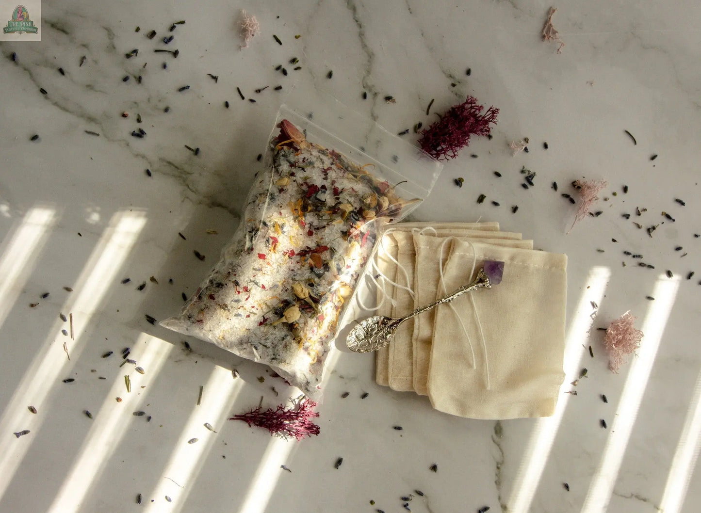 The Self Love bath tea set includes a clear bag of dried flower bath salts, empty muslin drawstring bags, an ornate spoon with a purple stone, and scattered petals, displayed on white marble with sunlight streaks.