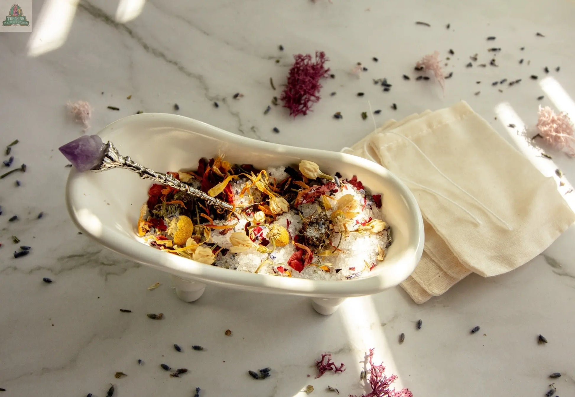The Self Love set features a white bathtub-shaped dish filled with dried flowers, herbs, bath tea, and salts, topped with a crystal wand—ideal for self-care rituals. Includes beige cloth bags and floral accents, displayed on marble.