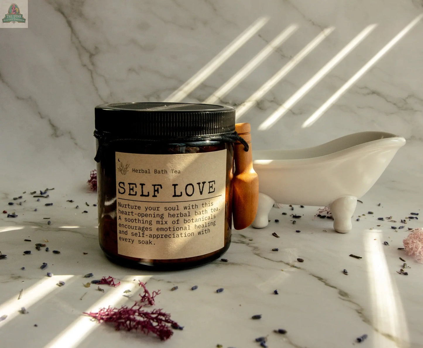 A jar of Self Love Herbal Bath Tea rests on marble with dried botanicals nearby. Infused with rose and vanilla essential oils, it’s perfect for your self-care ritual. A clawfoot tub and wooden spoon bask in sunlight.
