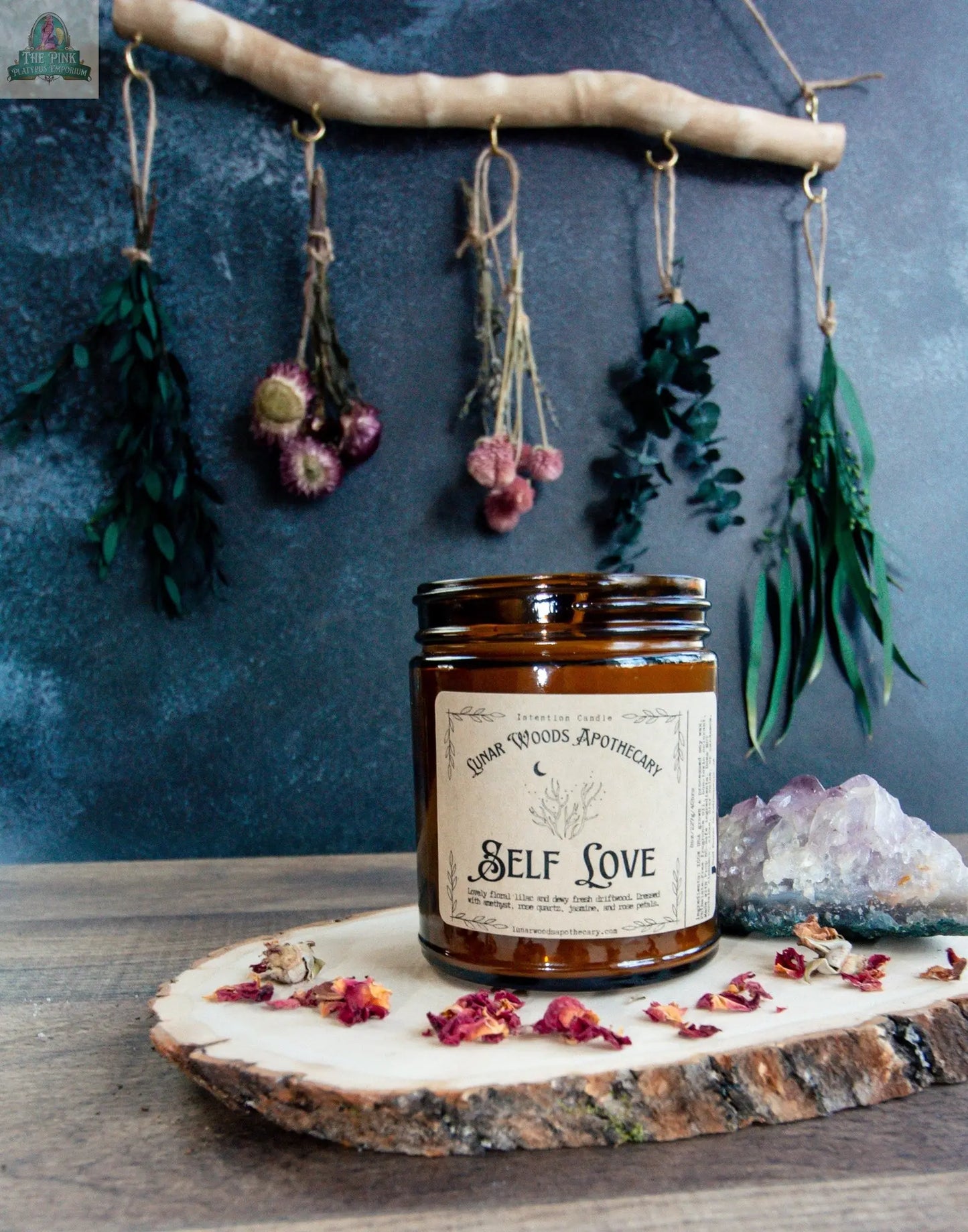 A brown glass Self Love candle sits on a wooden tray with dried flower petals next to an amethyst crystal, while dried flowers and plants hang on strings against a dark wall in the background.