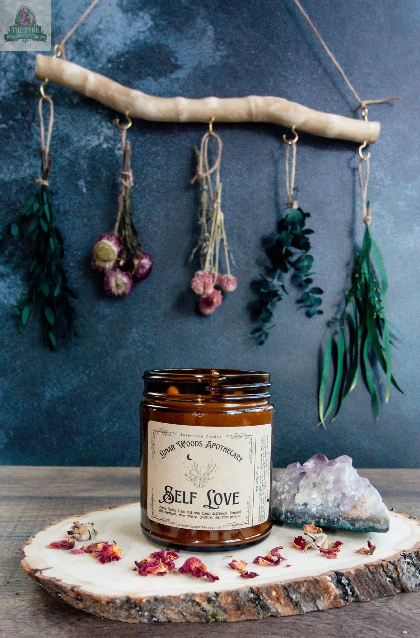 A brown glass "Self Love" candle sits on a wooden slab with dried petals, a rose quartz crystal, and a wall hanging of dried herbs and flowers, all set against a textured blue wall.