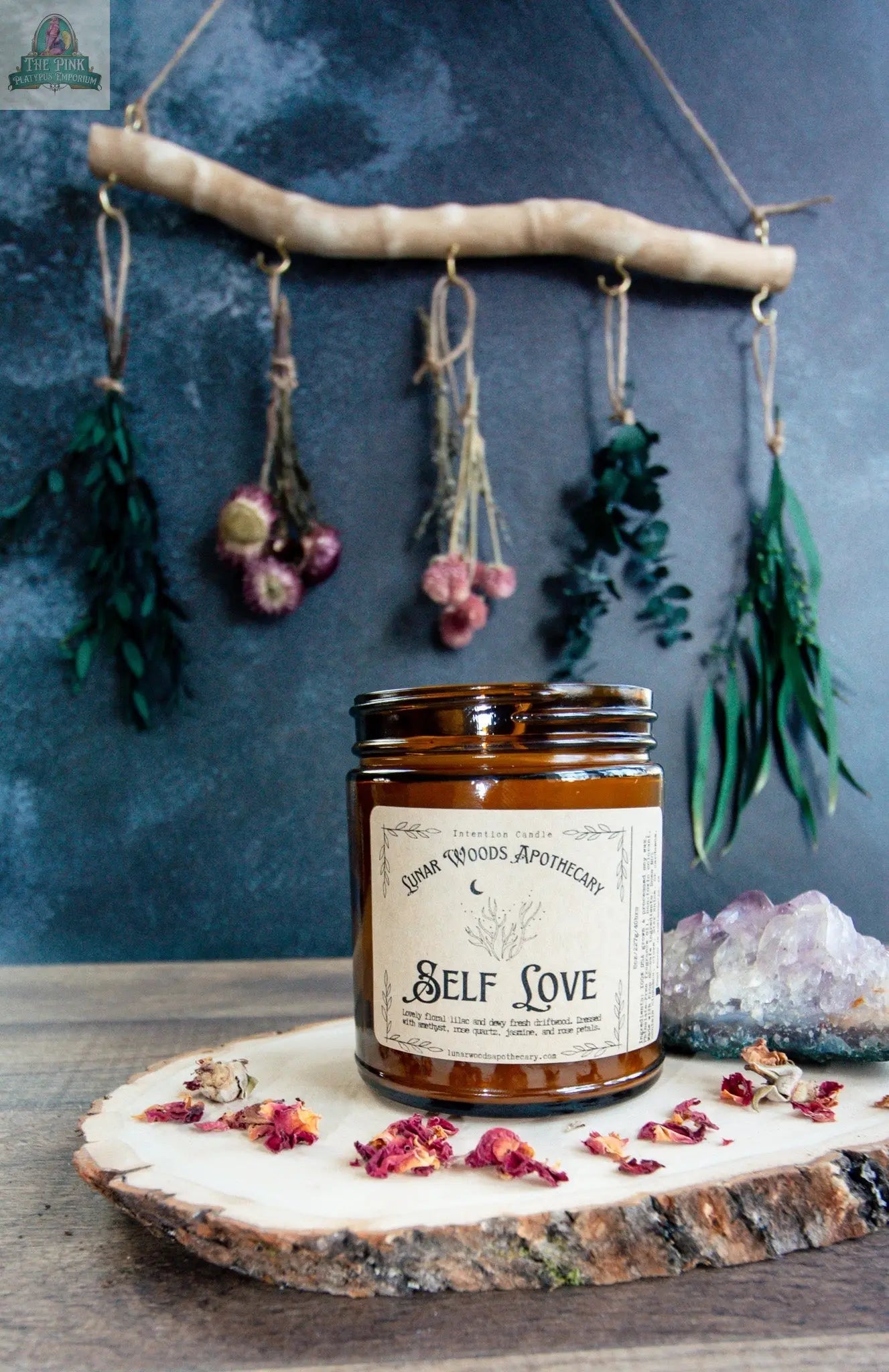 The Self Love candle in a brown glass jar sits on a wooden tray with dried rose petals. A rustic branch with dried flowers hangs behind it, and a rose quartz crystal cluster rests nearby on the tray.