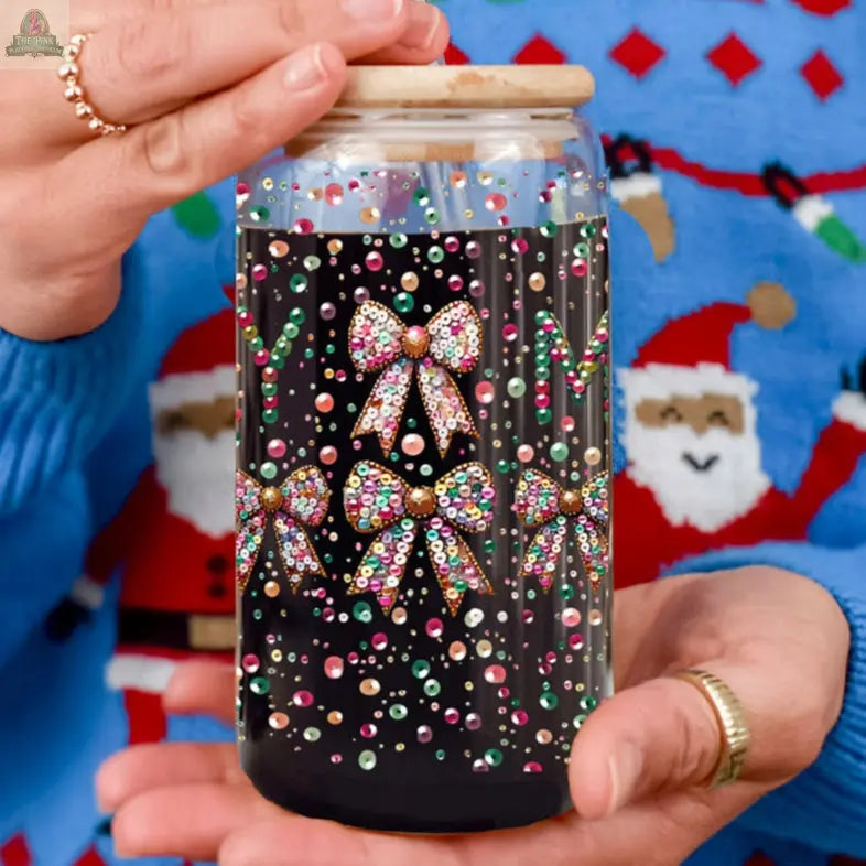 A person in a blue holiday sweater with Santa faces holds the Sequin MERRY tumbler featuring colorful, sparkling bows and dots, topped with a wooden lid. Gold rings adorn their fingers.