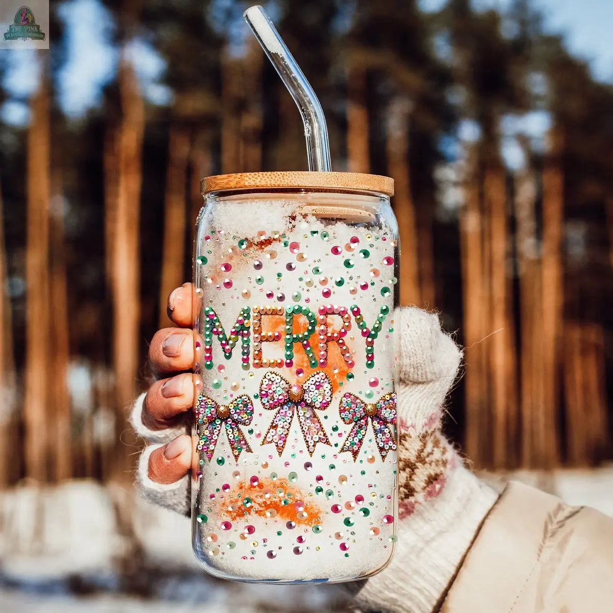 A gloved hand holds the Sequin MERRY tumbler, a festive holiday glass with a bamboo lid and sequin bow, filled with a snowy drink. Blurred trees and falling snow create a perfect seasonal backdrop.