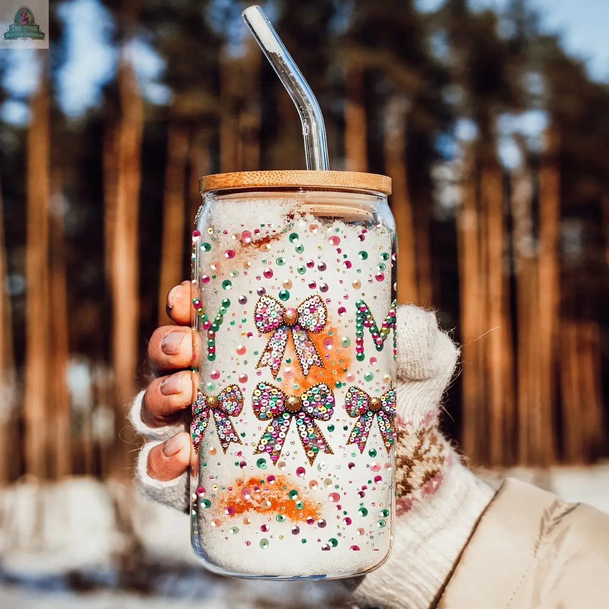 A hand in a fingerless glove holds the Sequin MERRY tumbler with a bamboo lid and straw, covered in colorful rhinestones, bows, and “MERRY,” against a snowy forest backdrop.