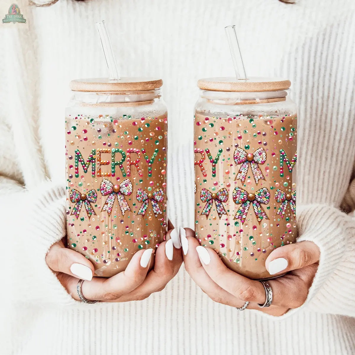 Two hands hold glass tumblers decorated with sequin MERRY, beads, and festive bows. The jars are filled with a brown drink and straws. The person wears a white sweater and matching white nail polish.
