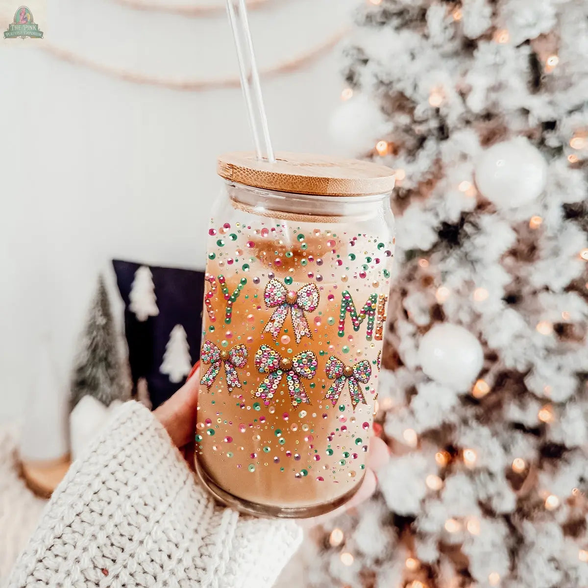 A hand in a cozy white sweater holds an iced coffee in a Sequin MERRY holiday tumbler adorned with festive bows and colorful sprinkles, with a blurred Christmas tree and ornaments in the background.