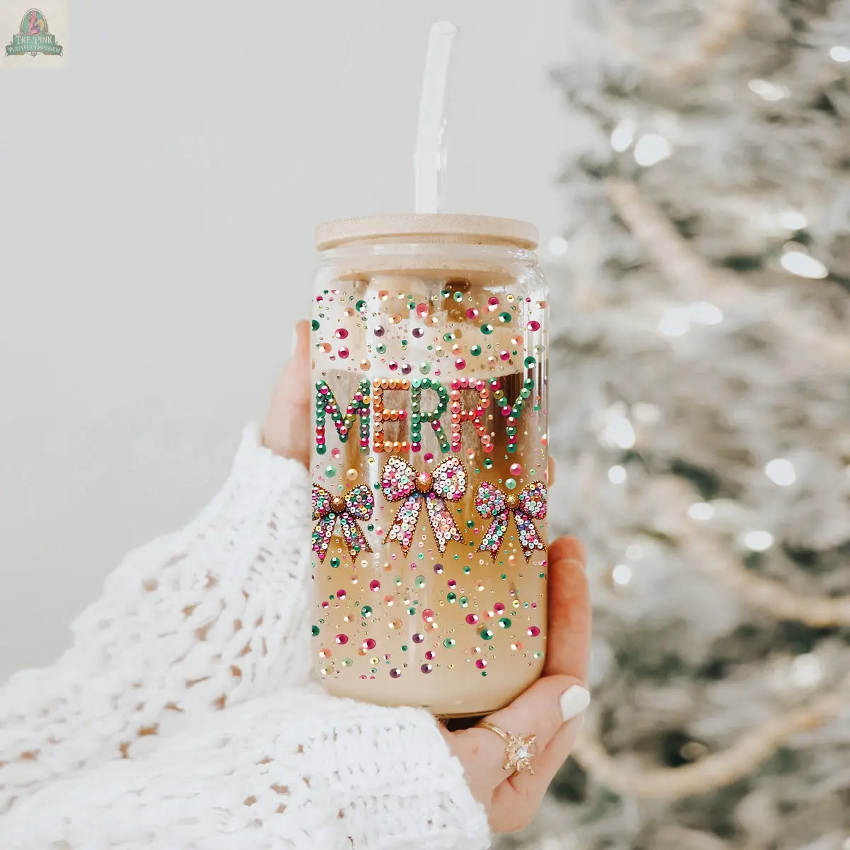 A hand holds the Sequin MERRY tumbler—a festive glass with a straw, colorful beads, bows, and "MERRY" lettering. A snowy Christmas tree glows in the background, adding to the holiday spirit.
