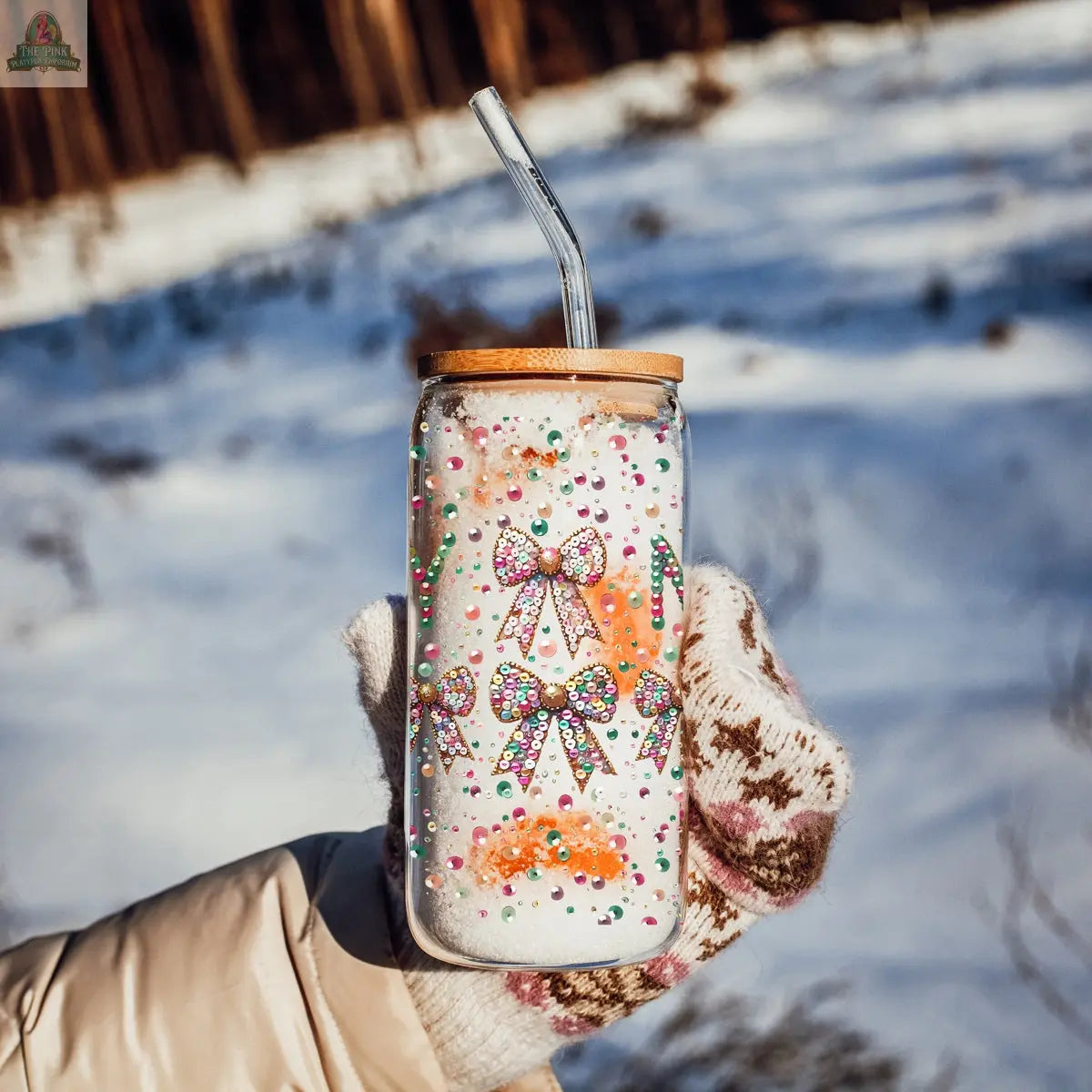 A hand in a patterned glove holds the Sequin MERRY tumbler with a metal lid and straw, decorated with colorful bows and confetti. Snowy ground and trees provide a festive, wintry backdrop.