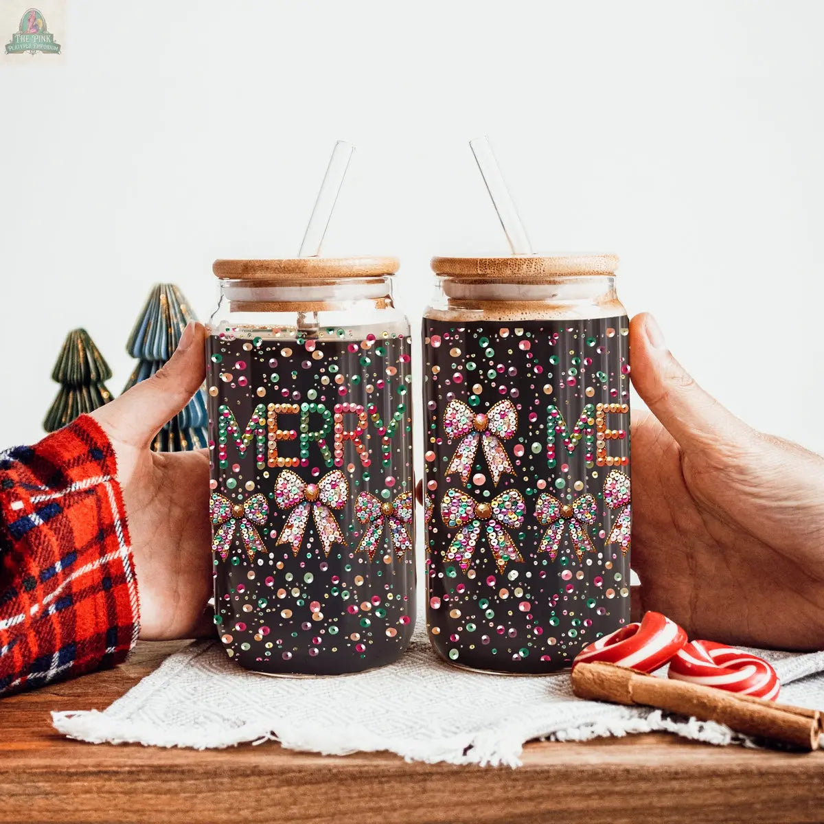 Two hands hold Sequin MERRY glass tumblers with bamboo lids and straws, featuring festive Merry text, sequin bows, and candy designs. Small Christmas ornaments and candy canes rest on a wooden table in the background.