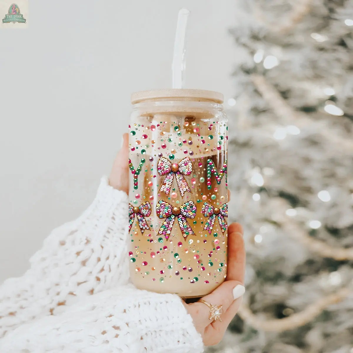 A hand in a white knitted sleeve holds the Sequin MERRY holiday glass tumbler filled with iced coffee, featuring colorful sprinkles and glittery bows, with a blurred Christmas tree in the background.