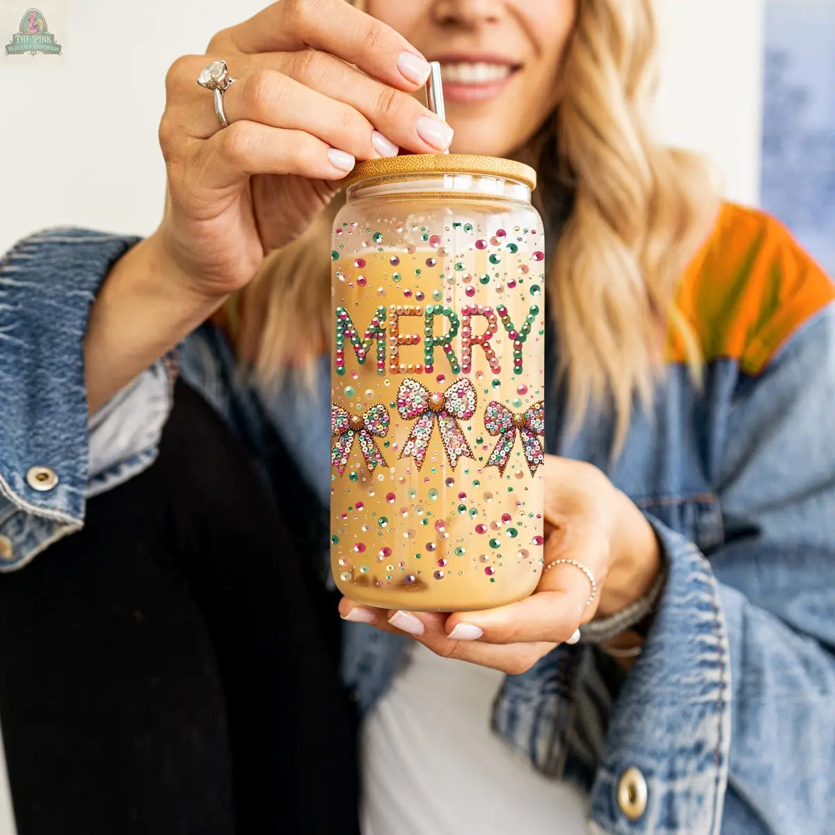 A woman in a denim jacket, wearing a ring, holds the Sequin MERRY tumbler filled with iced coffee. The holiday glass features colorful rhinestone bows, a metal straw, and a wooden lid.