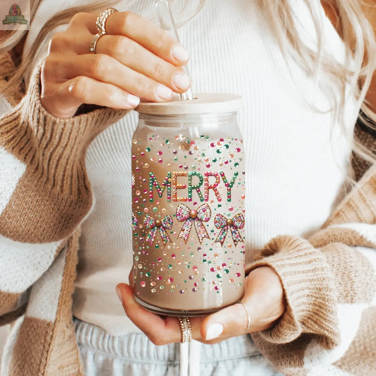 A woman in a white top and striped cardigan holds a Sequin MERRY holiday tumbler decorated with colorful rhinestones and bows, sipping her drink through a straw.