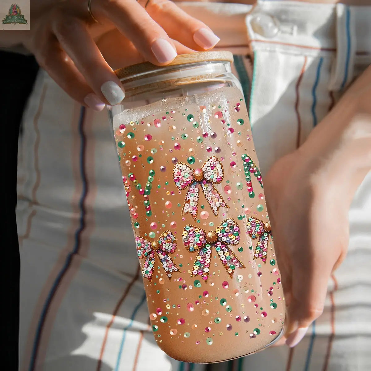 A person holds a Sequin MERRY tumbler filled with a light brown drink. The mason jar features colorful gem bows and dots, and the person is dressed in white pants with multicolored vertical stripes.