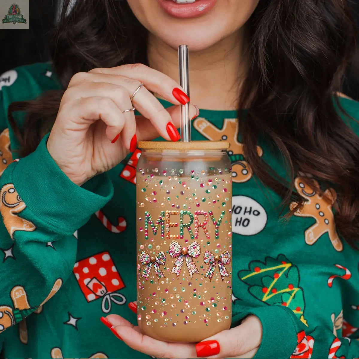 A woman in a festive green sweater with gingerbread and gift designs holds the Sequin MERRY tumbler glass jar, adorned with bows and colorful sprinkles, sipping her iced coffee through a metal straw.