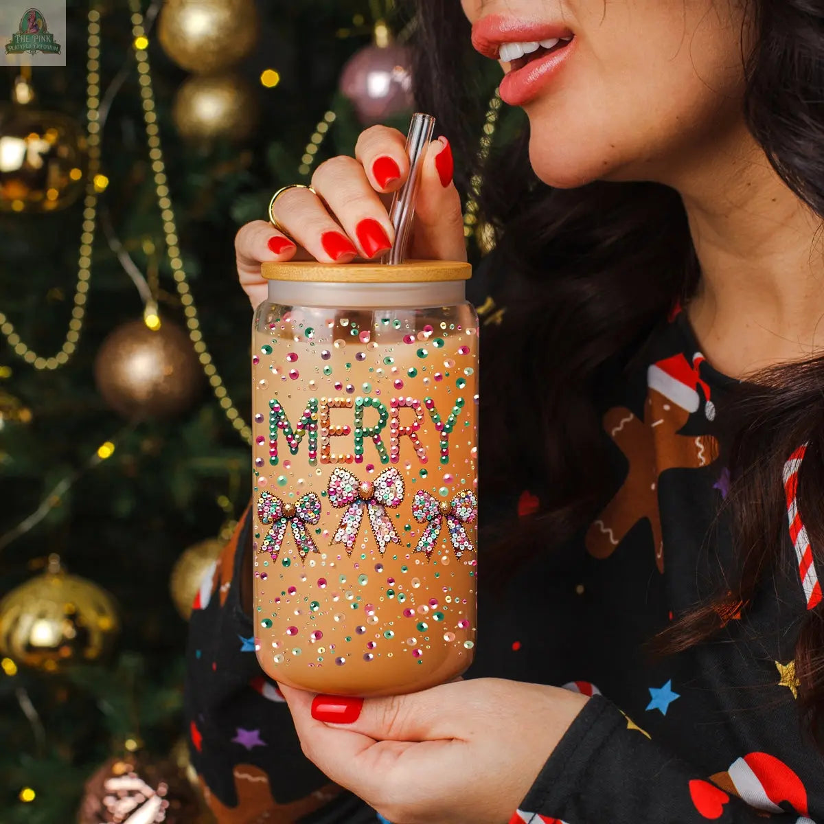 A woman with red nail polish sips from a Sequin MERRY tumbler glass jar with a metal straw. She wears a festive shirt, as a Christmas tree with gold ornaments sparkles in the background.