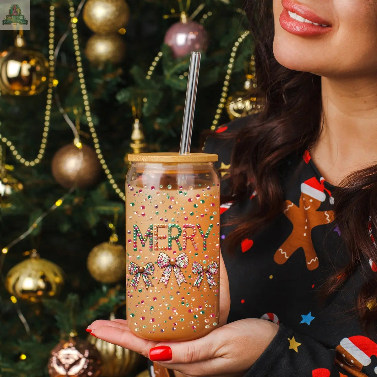 A woman in a festive sweater holds a Sequin MERRY tumbler with a straw and lid, decorated with bows. A sparkling Christmas tree with gold ornaments shines in the background.