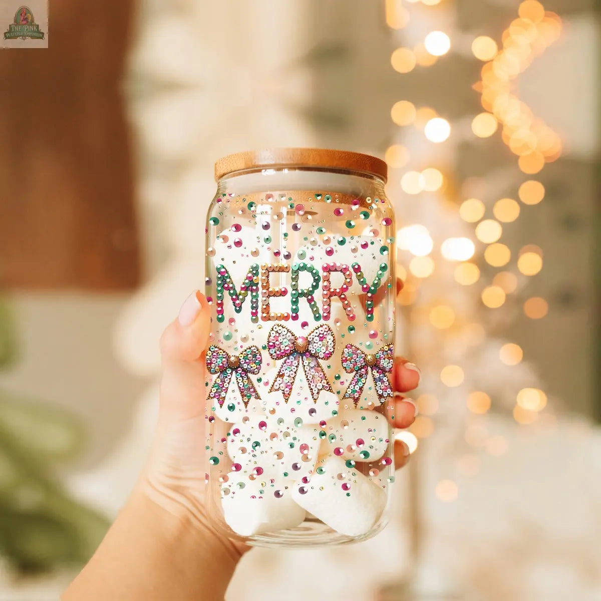 A hand holds a Sequin MERRY tumbler—a clear jar decorated with colorful beads spelling MERRY and three festive bows, filled with marshmallows. Blurred holiday lights and decor twinkle in the background.