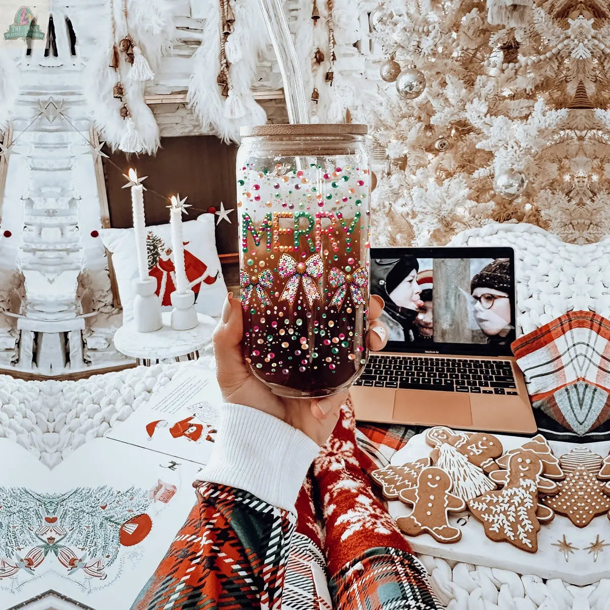 A hand in a festive sweater holds the Sequin MERRY tumbler jar reading “Merry Xmas” in a cozy Christmas setting with cookies, a laptop playing a holiday movie, coloring pages, and a softly glowing white Christmas tree.