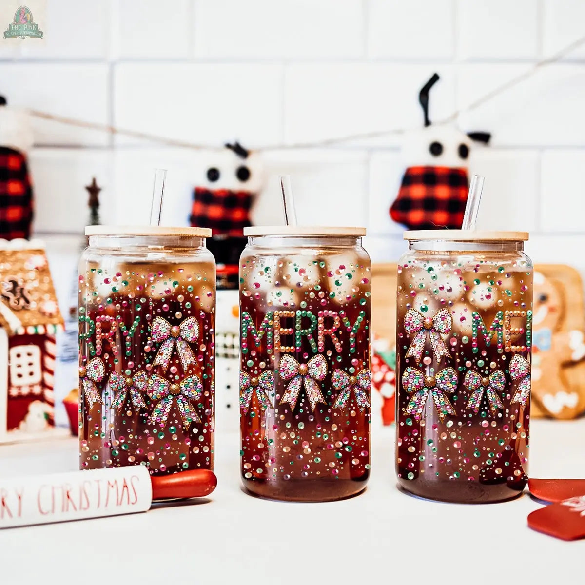 Three Sequin MERRY glass tumblers, each filled with a dark drink and topped with a colorful sequin bow, stand on a white tiled surface with holiday decor and stockings in the background, spreading festive cheer.