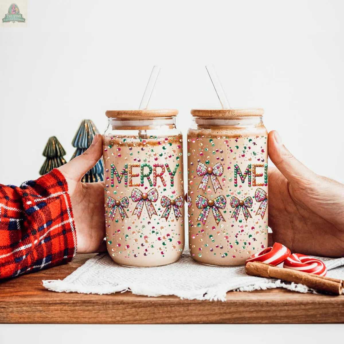 Two hands hold Sequin MERRY tumblers with festive straws, each glass decorated with colorful sprinkles and sequin bows. Mini Christmas trees and candy canes accent the background as the tumblers are filled with a creamy drink.