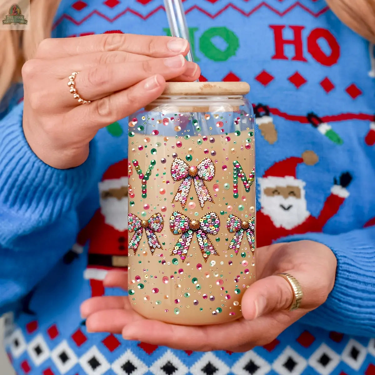 A person wears a festive blue Christmas sweater with Santa faces and holds the Sequin MERRY holiday glass tumbler, featuring a wooden lid, straw, and colorful, sparkling sequin bows and dots.