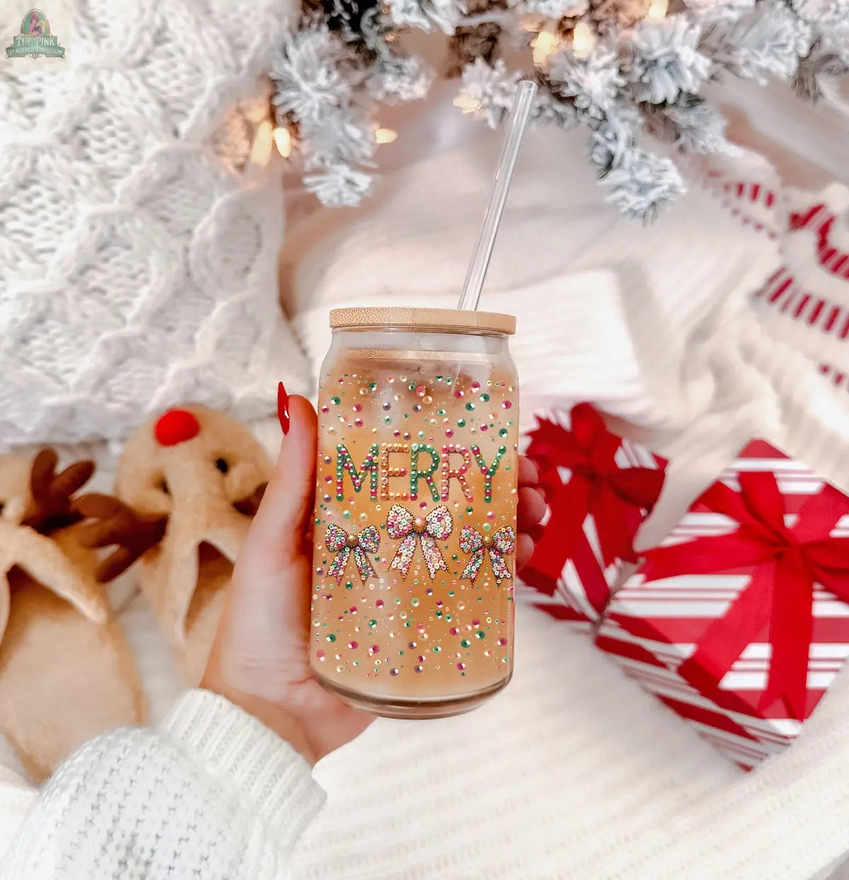 A hand holds the Sequin MERRY holiday glass tumbler topped with colorful sprinkles. In the background are a white knit blanket, reindeer slippers, snowy pine branches, and red-striped gift boxes.
