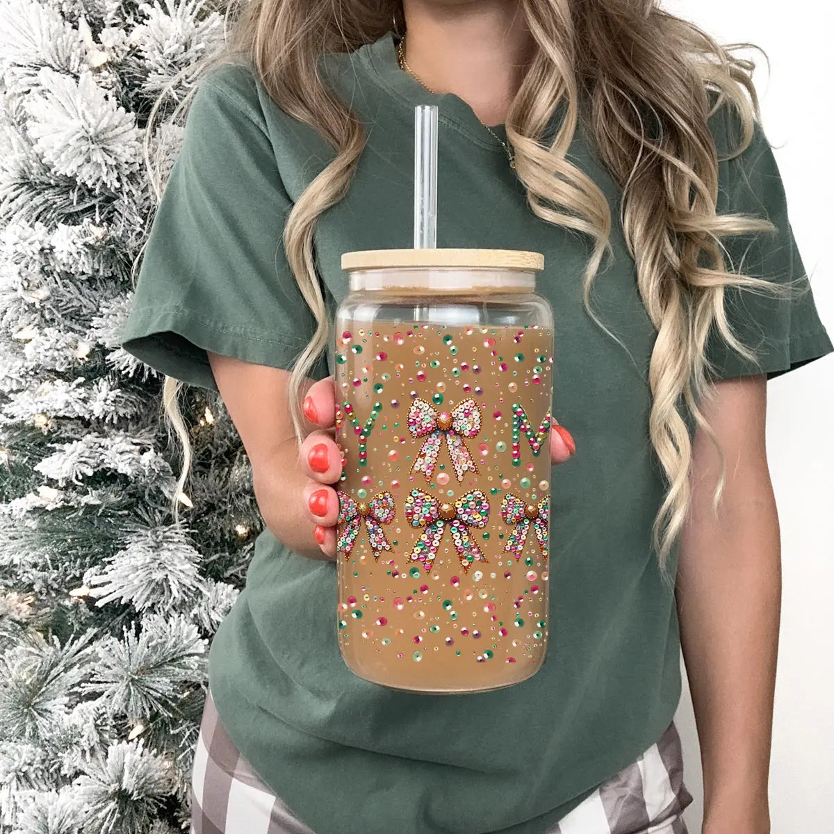 A woman in a green t-shirt holds the Sequin MERRY tumbler, decorated with festive bows and colorful sprinkles. The clear tumbler features a straw and bamboo lid, with a frosted Christmas tree softly glowing in the background.