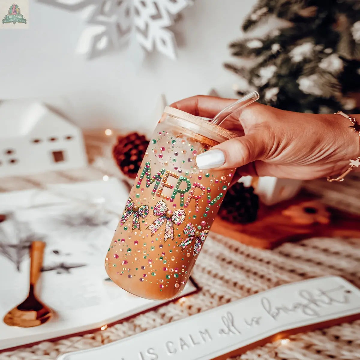 A hand holds a Sequin MERRY tumbler filled with iced coffee and decorated with colorful rhinestones, surrounded by festive holiday decor like a snowflake, pinecones, and a small Christmas tree.