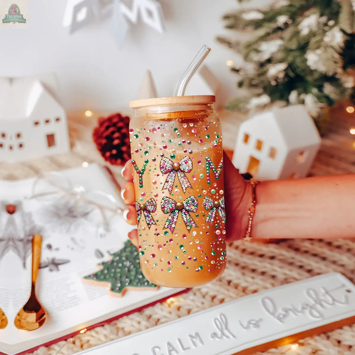 A hand holds the Sequin MERRY tumbler with lid and straw, decorated with colorful bows and dots, filled with an orange drink. Holiday decor, a tree, and an open book are in the background.