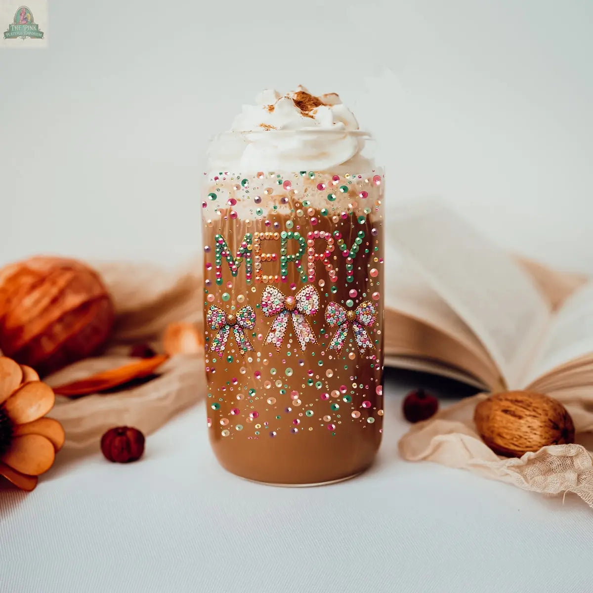 Celebrate in style with the Sequin MERRY glass tumbler—iced coffee with whipped cream, cinnamon, sprinkles, bows, and "MERRY." Surrounded by cozy fall accents like a pumpkin, nuts, and an open book, it’s perfect for festive gatherings.