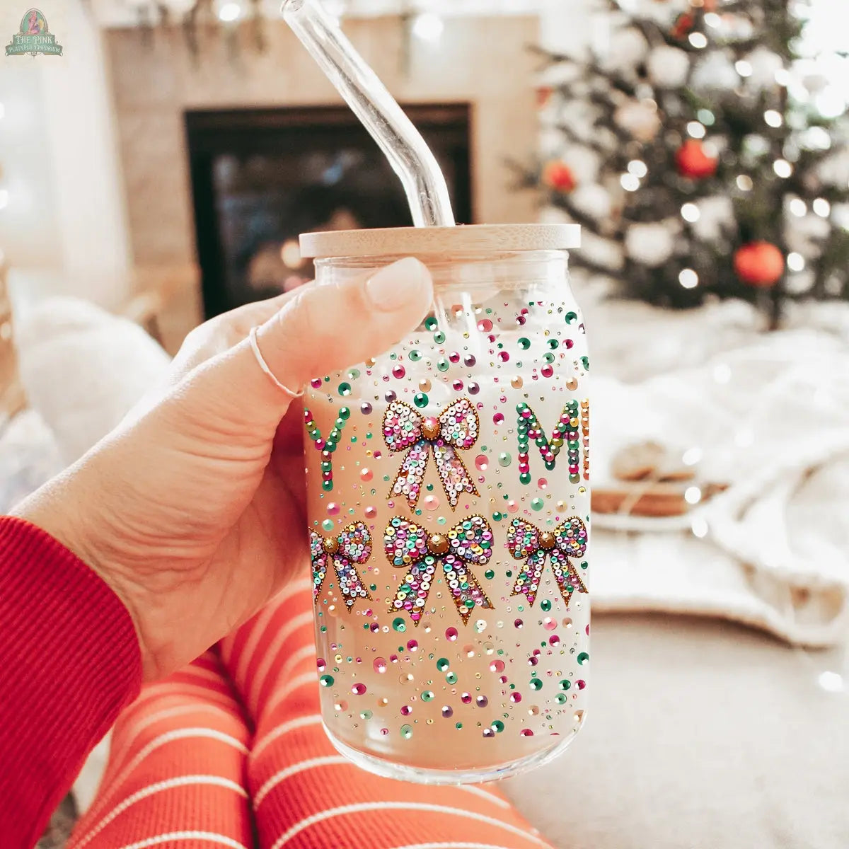 A hand in red striped pajamas holds a Sequin MERRY holiday glass tumbler with bamboo lid and straw, adorned with colorful bows and beads. A lit Christmas tree and fireplace blur in the background, creating cozy holiday vibes.