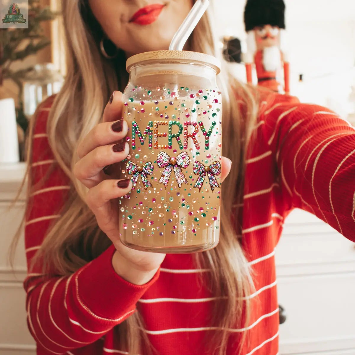 A woman in a red striped sweater holds the Sequin MERRY glass tumbler with a bamboo lid and straw, adorned with bows and "MERRY." The tumbler contains a light brown drink, while a nutcracker figure is visible in the background.