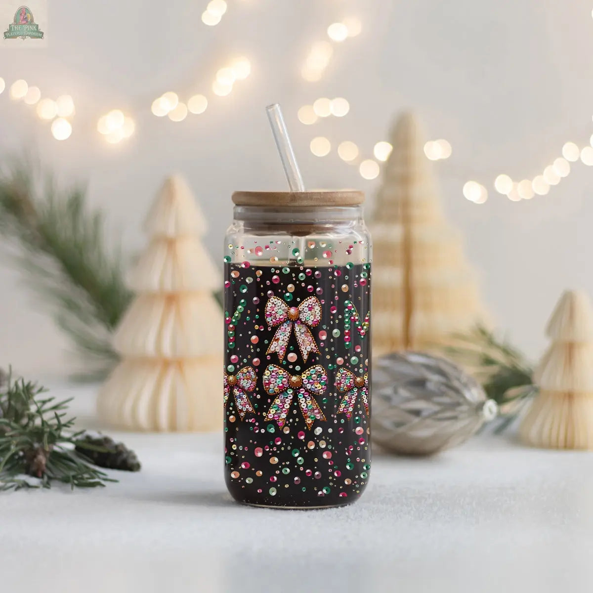 The Sequin MERRY glass tumbler with a wooden lid and straw, filled with a dark drink, is decorated with sequin bows and colorful dots. Festive ornaments and glowing fir branches in the background create a cozy holiday vibe.